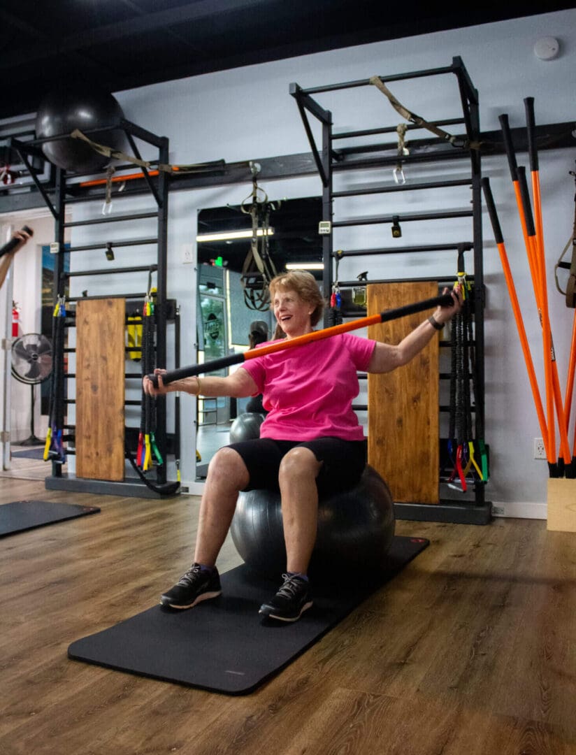 A lady holding a bar while sitting on a ball in the gym